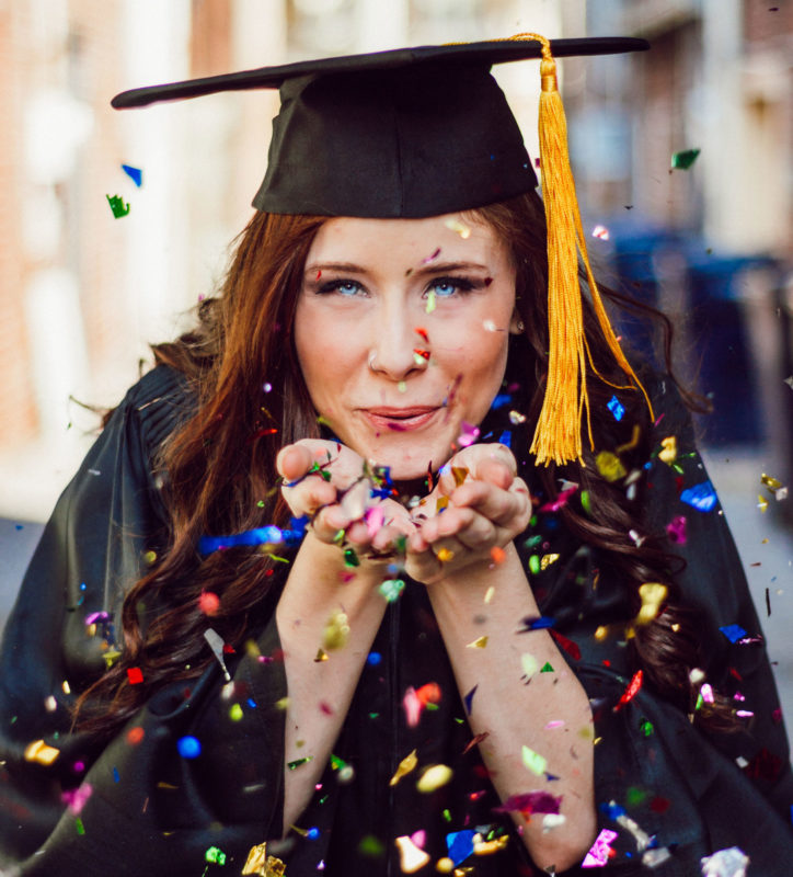 woman in graduation cap and gown blowing confetti from hands